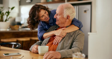 Smiling senior father and daughter embracing while enjoying tea at home. Elderly father and adult daughter bonding over tea and conversation. Warm moment between father and daughter sharing tea mugs while looking at each other.